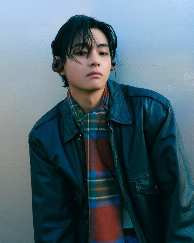 男性がイヤホンを着け、革のジャケットとチェック柄のマフラーを着て、壁に寄りかかっている姿。