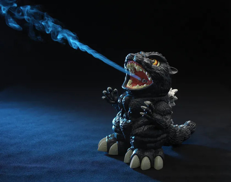 ゴジラのフィギュアが口から青い煙を吐いている様子を捉えた写真です。フィギュアは黒色で、白い爪と背びれが特徴的です。背景は暗く、青い光がフィギュアを照らしています。