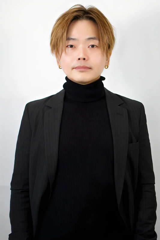 男性が正面を向き、黒いタートルネックセーターと黒いストライプのジャケットを着て立っています。背景は白です。