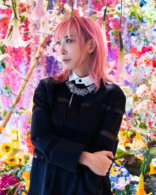 ピンク色の髪の女性が、花で飾られた背景の前に立っています。彼女は黒い服を着ており、胸元には装飾的なネックレスをしています。彼女はカメラから目をそらしており、やや遠くを見つめているようです。