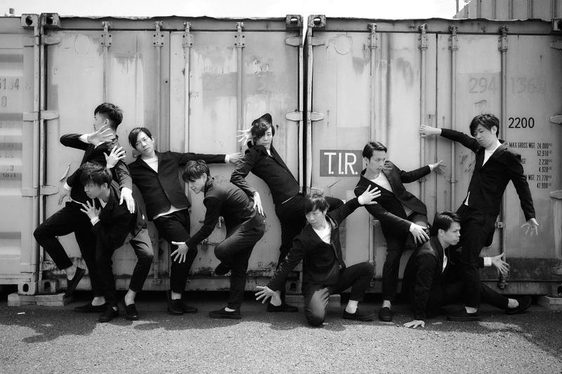 男性グループがコンテナの前でポーズをとっているモノクロ写真。メンバーは黒いスーツを着ており、さまざまなポーズをしています。