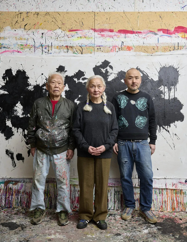 3人の人物が、絵の具が飛び散った壁を背景に立っている写真。左の男性は、赤いインナーと、絵の具で汚れたジャケットとズボンを着用している。中央の女性は、灰色のセーターと茶色のズボンを着用している。右の男性は、黒いセーターとジーンズを着用している。