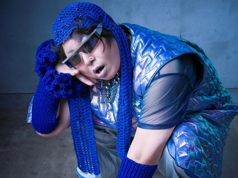 青色のニット帽、サングラス、青色のベストを着て、片手で顔を支えている人物のクローズアップ写真。背景は青色の壁。