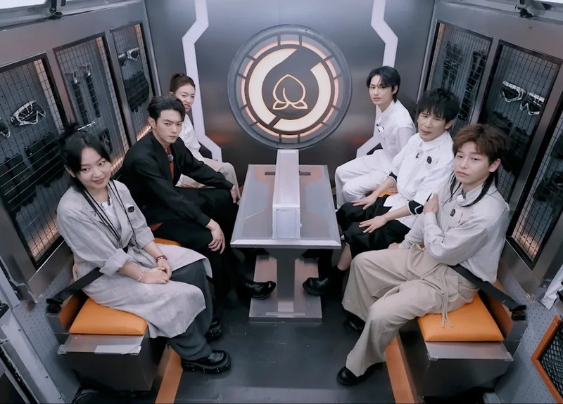 現代的で未来的な空間に6人の男女が座っている様子。彼らは中央のテーブルを囲んで向かい合い、何らかの会話や番組収録をしているように見える。壁には特徴的なエンブレムがある。