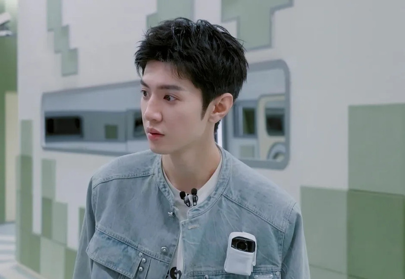 明るい背景の前で、デニムジャケットを着用した若いアジア人男性のバストアップショット。マイクと小型カメラを身につけ、横を向いて何かを見つめている。