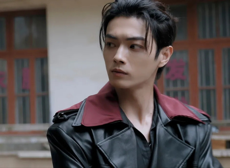 黒いレザージャケットと赤い襟が特徴的な若い男性のポートレート。彼は真剣な表情で視線を横に向けており、背景には窓のある建物と赤い文字が見える。スタイリッシュで落ち着いた雰囲気の人物像。