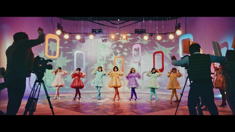 ステージでポーズをとるカラフルな衣装の女性アイドルグループ。撮影クルーも映っている。