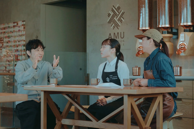 3人の人々がテーブルを囲んで話している様子。中央の女性はエプロンを着けており、左側の男性は両手を広げて話している。背景にはコーヒー豆の容器やロゴが見える。