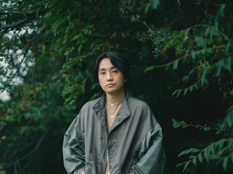 木々を背景に、ジャケットを着た男性が正面を見ているポートレート写真です。