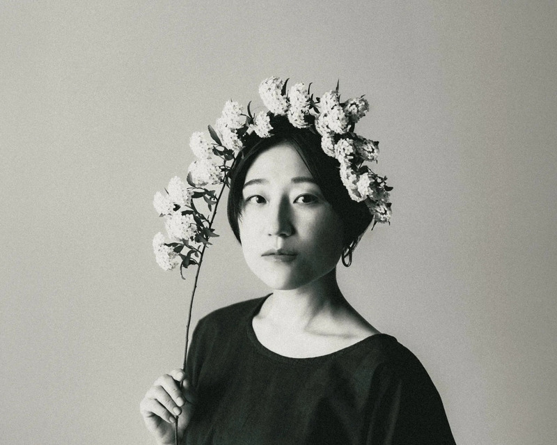 花冠をつけた女性のモノクロポートレート。女性は、白い花が咲いた枝を持ち、カメラ目線で少し悲しげな表情をしています。