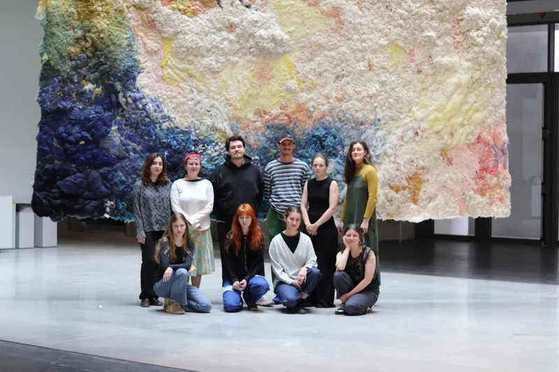 大きなテキスタイル作品の前で記念撮影をする人々の写真。作品は様々な色合いの繊維で構成され、壁に掛けられている。人々は様々な年齢層で、カジュアルな服装をしている。
