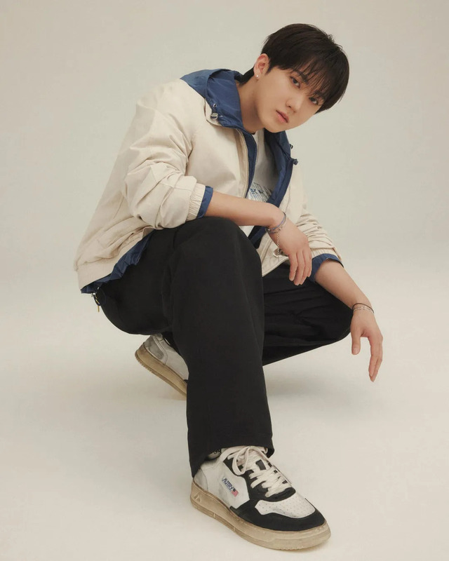 男性がしゃがみこんでカメラを見ている写真。彼は白いジャケットと黒いズボンを着ており、足元にはスニーカーを履いている。背景は白。