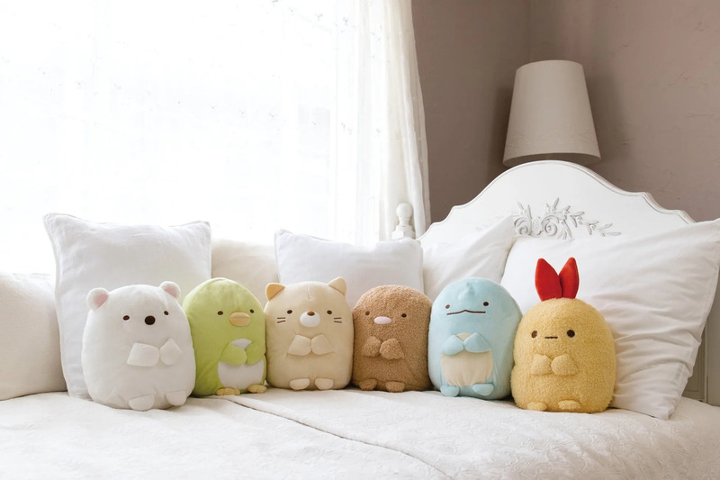白いベッドの上に、すみっコぐらしのぬいぐるみたちが並んで置かれている様子を写した画像。それぞれのキャラクターが可愛らしく、様々な色と形で表現されている。背景には白いカーテンと枕があり、柔らかい雰囲気を作り出している。