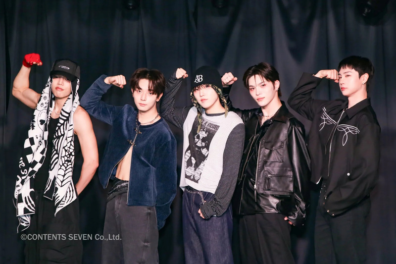 5人の男性がポーズをとってカメラに向かって立っている写真。彼らは皆、さまざまな衣装を着ており、黒い背景の前に立っています。