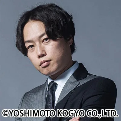 スーツを着た男性のポートレート写真です。彼はカメラ目線で、暗い背景に立っています。