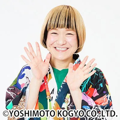 女性が両手を広げて笑顔で写っている写真です。彼女は明るい髪型をしており、カラフルな服を着ています。