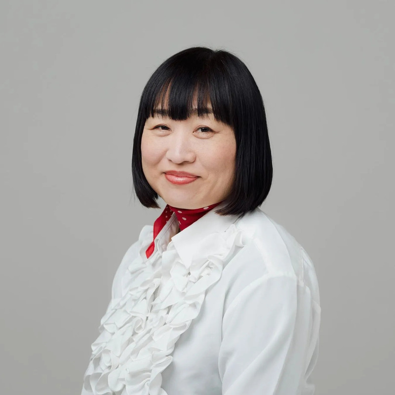 白いフリルシャツを着て、赤いスカーフを首に巻いた、黒髪の女性のポートレート。彼女はカメラに向かって微笑んでいます。