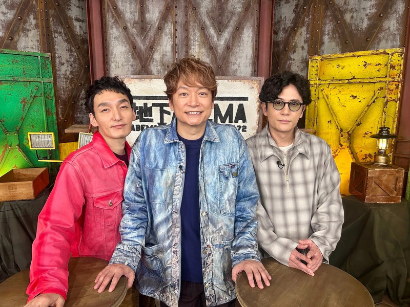 3人の男性が写っている写真。真ん中の男性はデニムのジャケットを着ており、他の2人はそれぞれ赤いジャケットとチェックのシャツを着ている。背景には、古びた看板や扉が写っている。