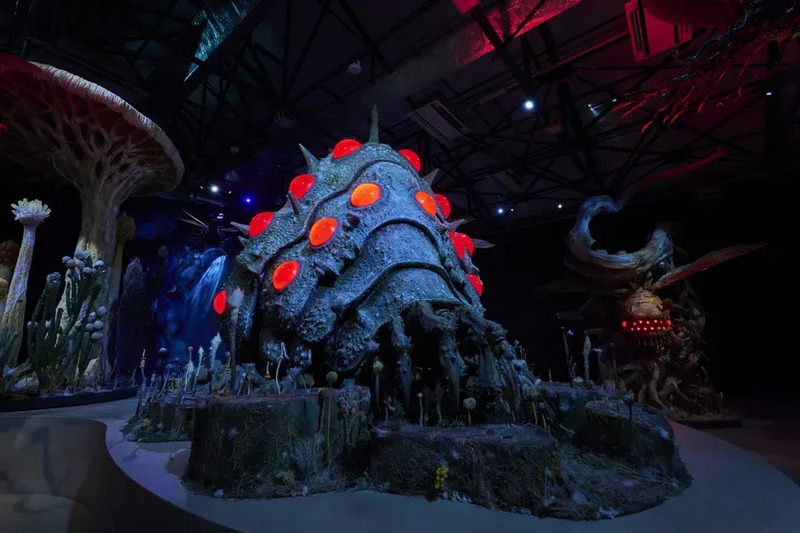 暗い空間に展示されている、巨大な生物の彫刻とキノコのような造形物。赤く光る目のようなものが特徴的。
