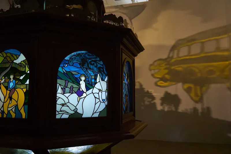 ステンドグラスの展示。中央には2つの窓があり、それぞれ異なるアニメのシーンが描かれている。左側の窓には、女性が黄色い動物に乗っている様子が描かれ、右側の窓には、少女が白い動物に乗っている様子が描かれている。壁にはバスの影が映し出されている。