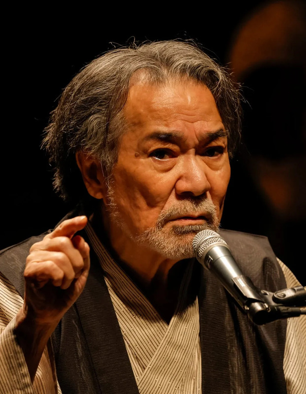 マイクに向かって話す男性のクローズアップ。彼は指を指し、険しい表情で話している。