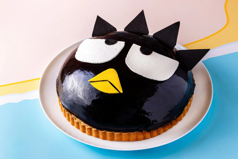 バッドばつ丸の顔を模したケーキ。黒いケーキの表面には、白い目と黄色のくちばし、黒い三角の耳が施されている。ケーキは白い皿に載せられ、背景は水色とピンク色。