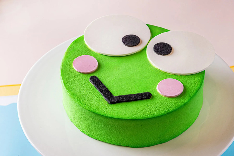 カエルの顔を模したケーキが白い皿の上に置かれています。ケーキは緑色で、白い丸い目、黒い瞳、ピンクの頬、黒い口が特徴です。