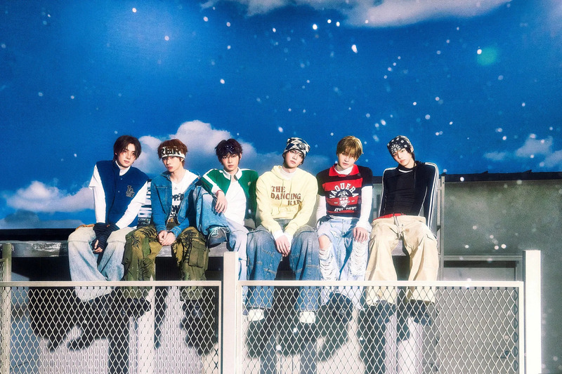 6人の男性が空を背景に座っている写真。彼らはカジュアルな服装をしており、それぞれ異なるスタイルをしています。背景には星がちりばめられた青い空があります。