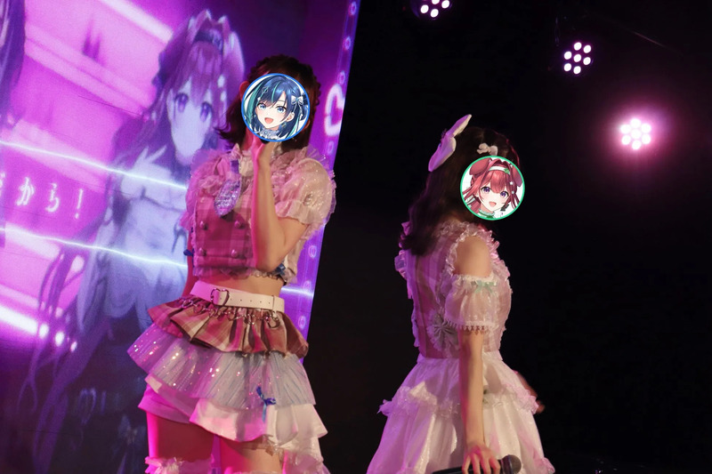 ステージでパフォーマンスをする2人の女性アイドル。ピンク色の照明の中で、彼女たちはそれぞれの衣装を着ており、顔にはアニメのキャラクターが描かれています。