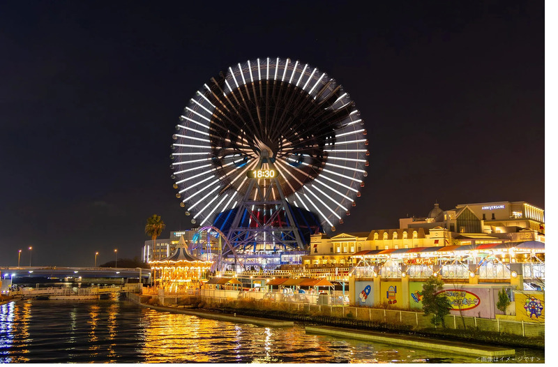 夜の観覧車と周辺の建物の写真。観覧車には18:30の時刻が表示され、ライトアップされている。水面に光が反射し、遊園地のような雰囲気。