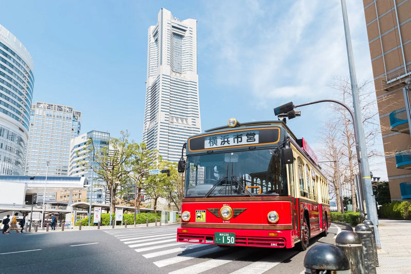横浜の街並みを走る赤いバスの鮮やかな写真です。バスは「あかいくつ」と呼ばれ、観光客に人気のようです。背景には高層ビルがそびえ立ち、都市の活気を感じさせます。