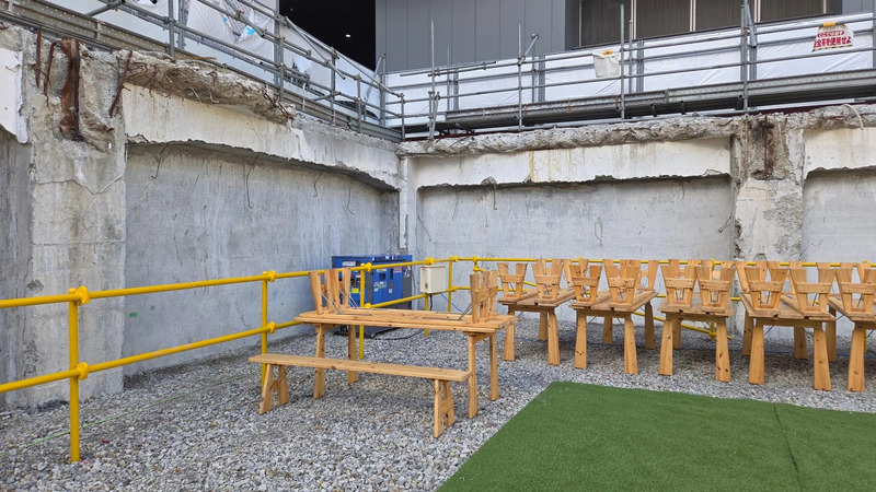 建設現場と思われる場所で、木製のテーブルと椅子が置かれています。壁はコンクリートで覆われ、足場が組まれています。