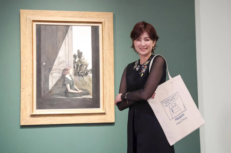 女性が絵画の前に立っており、彼女は白いトートバッグを持っており、そのバッグには「ピーナッツ」のキャラクターと「Andrew Wyeth」というテキストが描かれています。絵画は、ドアの前に座っている女性が描かれています。
