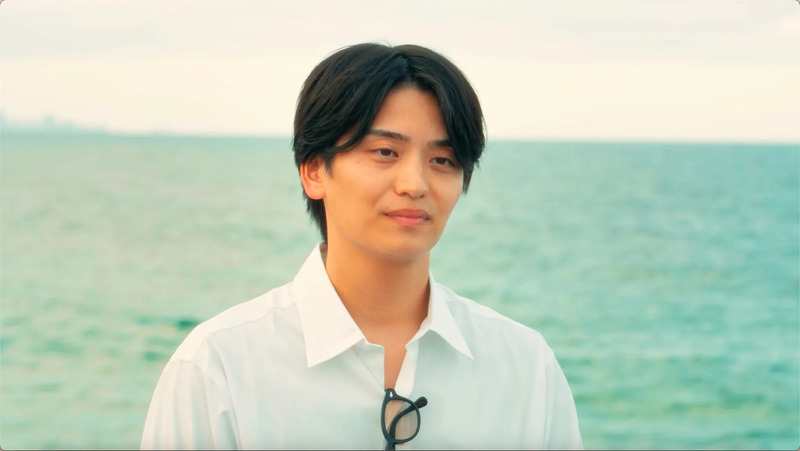 男性が海を背景に立っている写真です。彼は白いシャツを着ており、少し微笑んでいます。彼の首にはメガネがぶら下がっています。