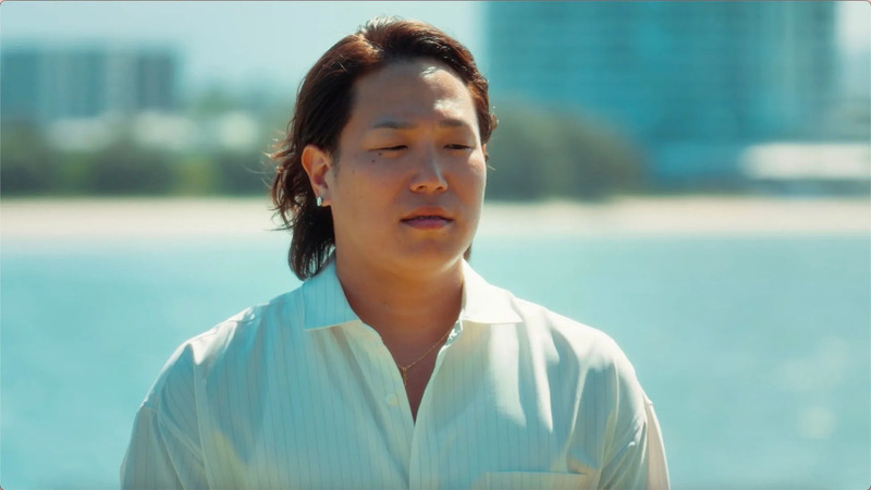 男性が屋外でポーズをとっている様子。彼は白いシャツを着ており、海を背景にしています。