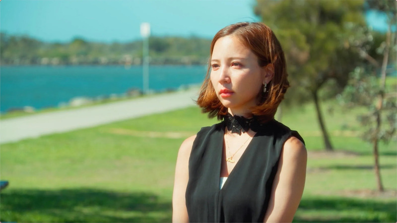 公園にいる女性のポートレート。女性は黒い服を着ており、首にはレースのチョーカーとネックレスを着けている。背景には水辺と緑の芝生が見える。