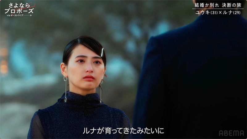 女性が涙を浮かべている様子。背景には男性の背中が見え、ドラマのワンシーンを切り取ったような画像です。