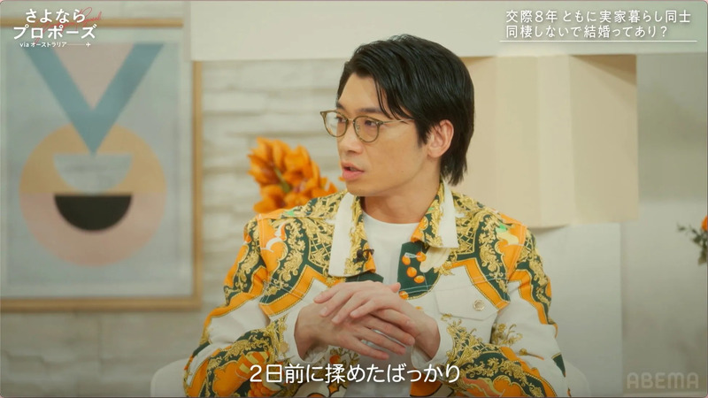 番組のセットで話す男性。番組タイトルと、交際期間や結婚に関するテキストが表示されている。