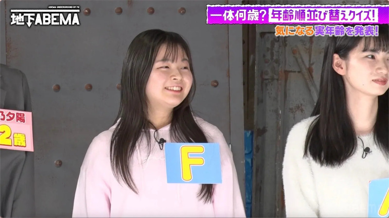 女性が出演している日本のテレビ番組のワンシーン。出演者は、ピンクの服を着た女性と、白いセーターを着た女性の2名。女性たちの前には、それぞれ「F」と書かれた札と、年齢を示唆する札がある。番組のロゴやタイトルも確認できる。