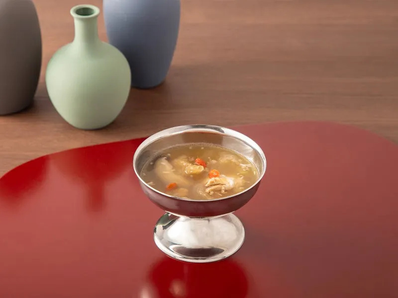 銀色の器に入ったスープのクローズアップ。背景には、3つの花瓶と赤いテーブルマットがある。