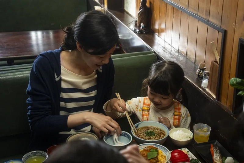 母親と子供が食事をしている様子。子供は箸を使って麺類を食べようとしており、母親はそれを見守っている。食事には麺類、ご飯、おかずなどが含まれている。