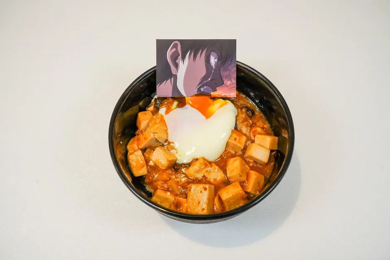 麻婆豆腐丼の上に温泉卵が乗った料理の写真です。黒い器に入っており、アニメのキャラクターが描かれたカードが添えられています。